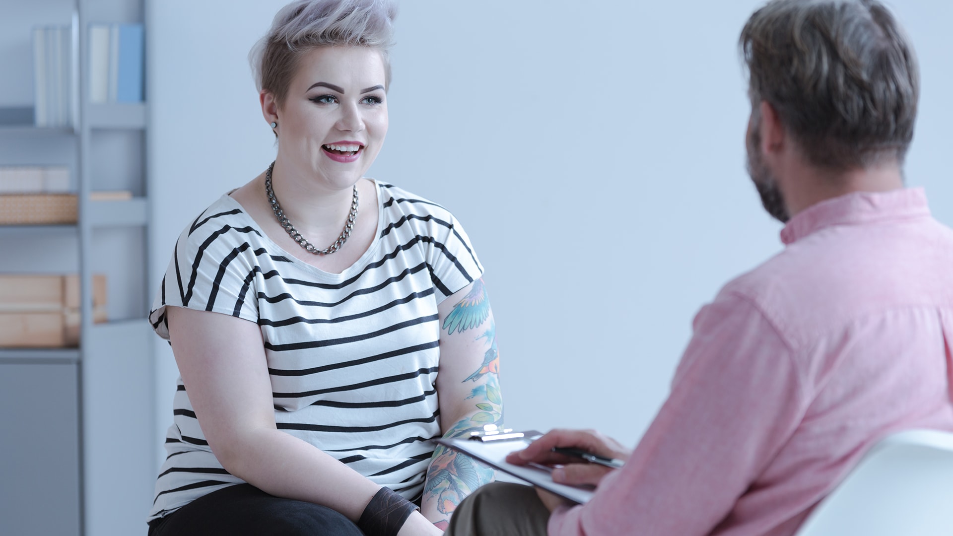 Client with short hair and tattoos smiling while talking with a health professional holding a clipboard